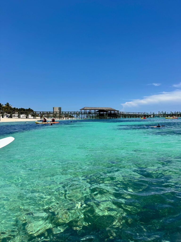 Turquoise water near a sandy beach and a wooden pier with a thatched shelter; people kayaking nearby. Grand hyatt baha mar